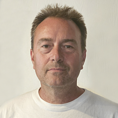 A portrait of Faren Cooper, a man with short brown hair and light stubble, wearing a plain white t-shirt, posed against a neutral light-colored background.