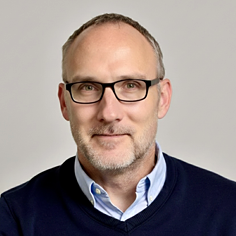 A headshot of James K. Lawton, a man with short hair and glasses, smiling slightly and wearing a blue sweater over a light blue collared shirt.