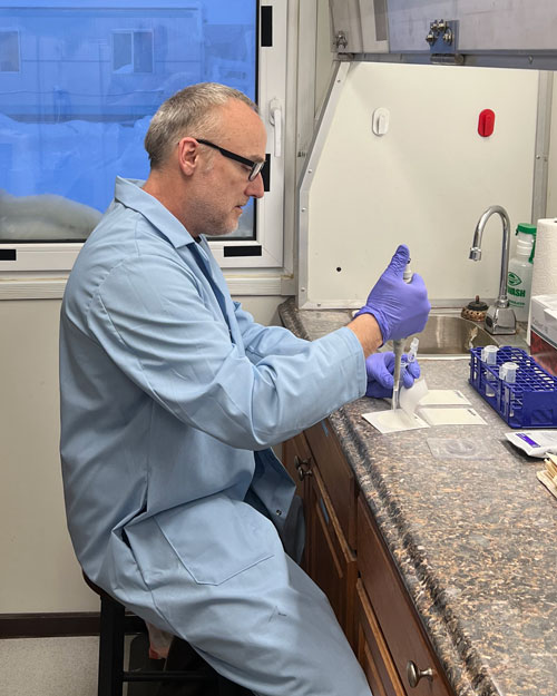 A man in a light blue lab coat and glasses sits on a stool at a lab bench. He is wearing purple gloves and using a pipette to transfer liquid into small tubes. A window and a sink are visible in the background.