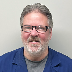 A headshot of Lance W. Morris, a smiling middle-aged man with graying hair and a beard, wearing black-rimmed glasses and a blue button-down shirt.