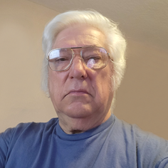 A headshot of Ralph E. Allphin, a man with white hair and large aviator-style glasses, wearing a blue t-shirt.