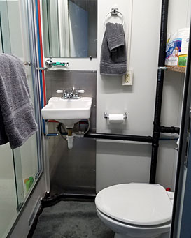 A compact, modern bathroom inside a modular unit featuring a white pedestal sink, a toilet, a glass-door shower, and exposed black plumbing pipes against white walls.
