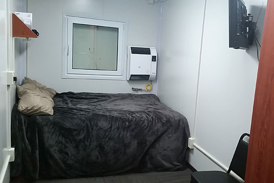 A small, functional bedroom with white walls, featuring a bed with a dark grey blanket, a window, a wall-mounted heater, and a television.