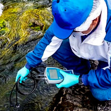 Environmental technician in a blue hat and protective gloves using a portable water quality meter to test a rocky shoreline stream.