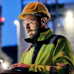 Close-up of a bearded environmental technician in a yellow hard hat and high-visibility safety jacket operating a laptop outdoors at night.