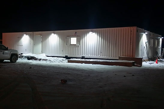 An exterior night shot of a long, white modular facility comprised of joined shipping containers, illuminated by several wall-mounted security lights, with snow covering the surrounding ground.