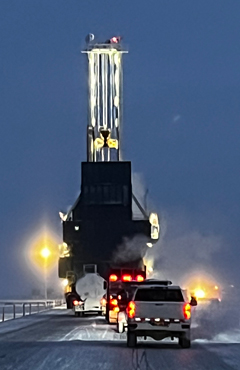 A white pickup truck following a massive industrial drilling rig being transported down a snow-covered Arctic road at twilight.