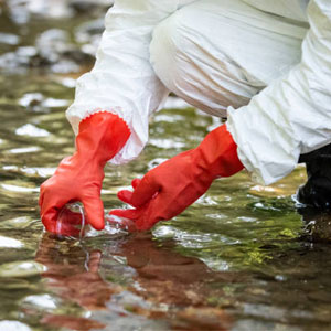 Close-up of an environmental technician in a white protective suit and red rubber gloves collecting a water sample from a shallow stream using a glass jar.